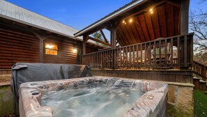 A hot tub in front of a wooden cabin with a deck and a stone wall. - Lazy J Hideaway -Hot Tub, fire pit, secluded cabin (Broken Bow)