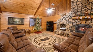 A log cabin interior with leather sofas, a stone fireplace, a Christmas tree, and a ceiling fan. - Lazy J Hideaway -Hot Tub, fire pit, secluded cabin (Broken Bow)