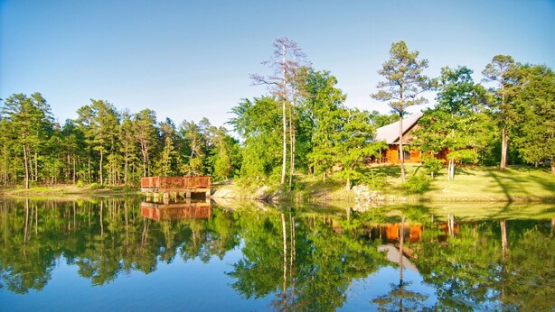 A lakeside cabin with a dock extending into the water, surrounded by dense trees and a clear sky. - Lazy J Hideaway -Hot Tub, fire pit, secluded cabin (Broken Bow)