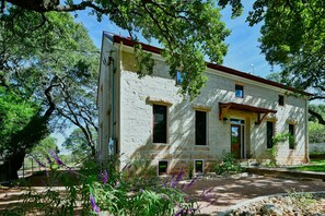 A two-story stone building with a red roof, surrounded by trees and purple flowers.