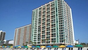 A high-rise hotel with multiple balconies, located on a beach with umbrellas and beach chairs.