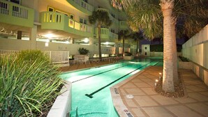 An indoor swimming pool with a tiled deck, surrounded by palm trees and a multi-story building with balconies.