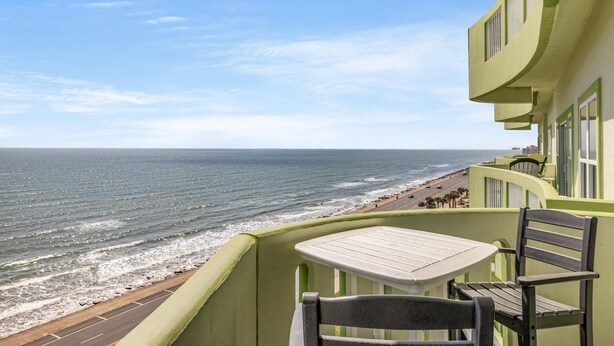 A balcony with a table and chairs overlooking a beach with a road and buildings in the distance.