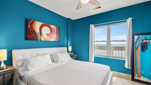 A bedroom with a large bed, a ceiling fan, a window with curtains, and a painting on the wall.