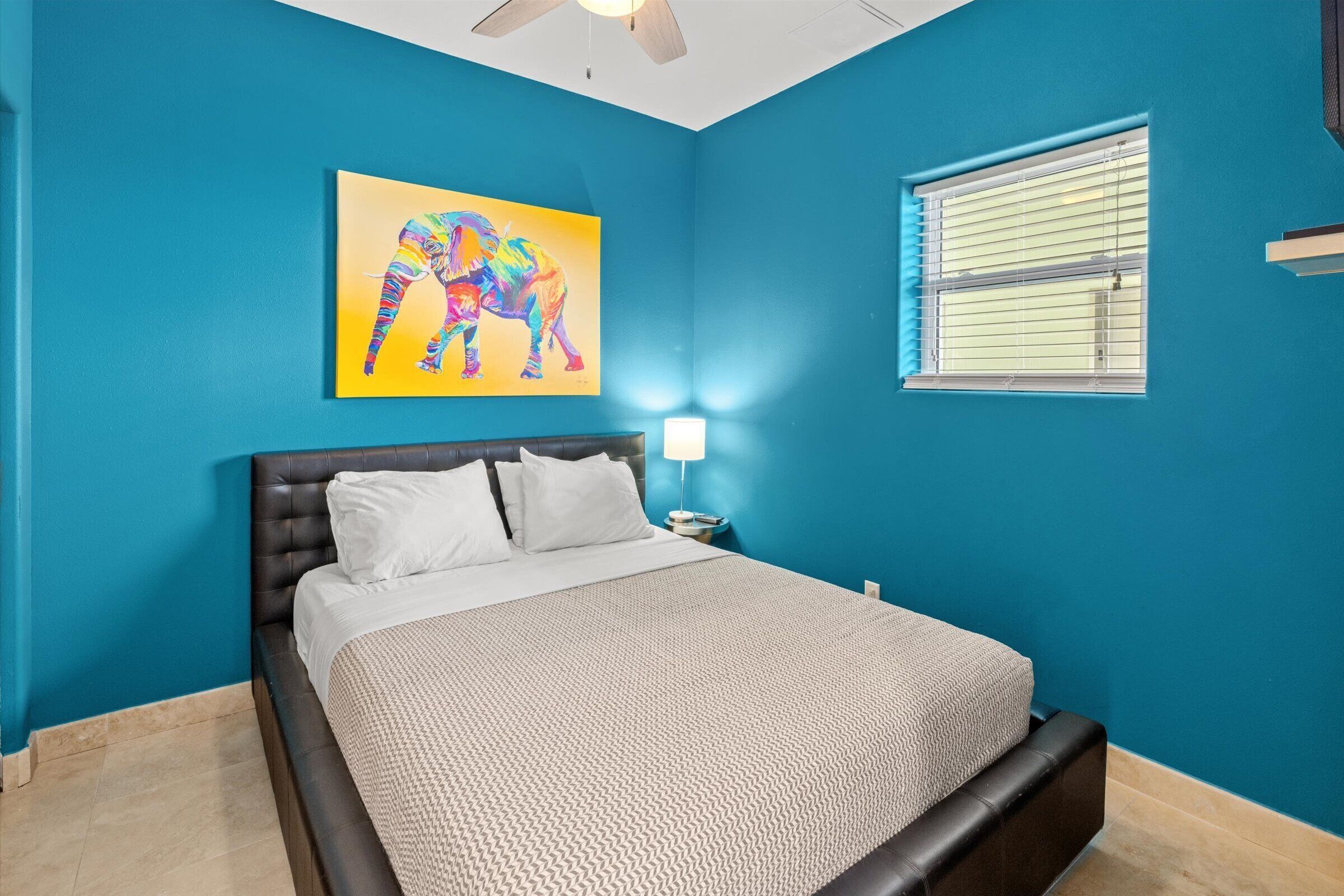 A bedroom with a bed, a nightstand, a lamp, a window with blinds, and a ceiling fan.