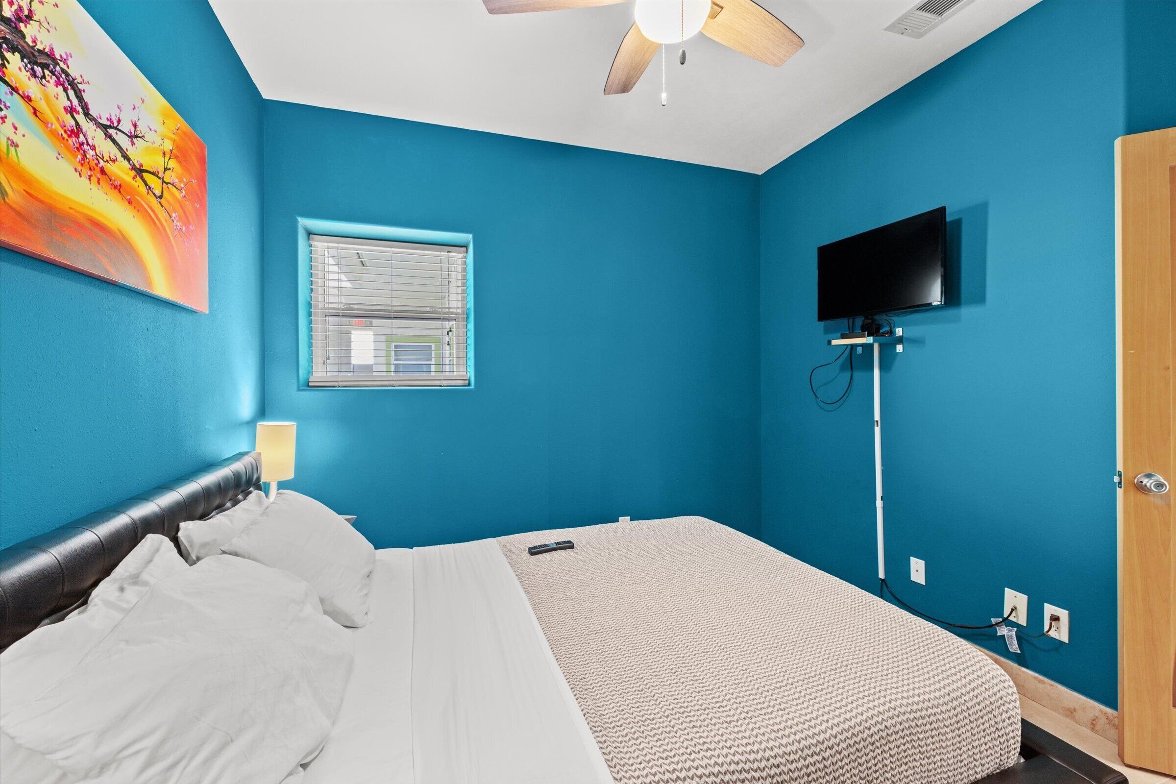 Een slaapkamer met een bed, een plafondventilator, een aan de muur bevestigde televisie en een raam met jaloezieën.