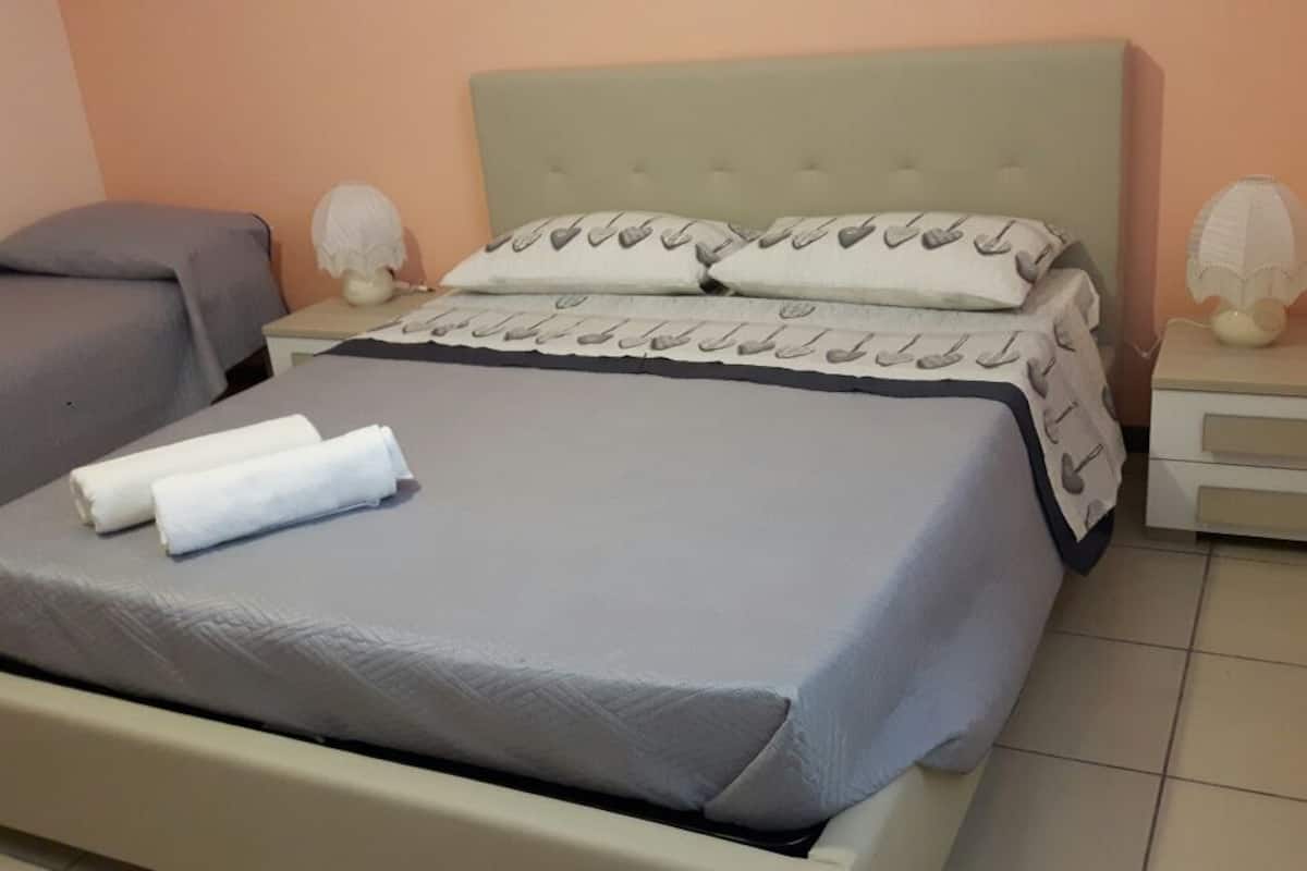 A single bed with a grey bedspread and white pillows, a bedside table with a lamp, and a nightstand with a lamp.