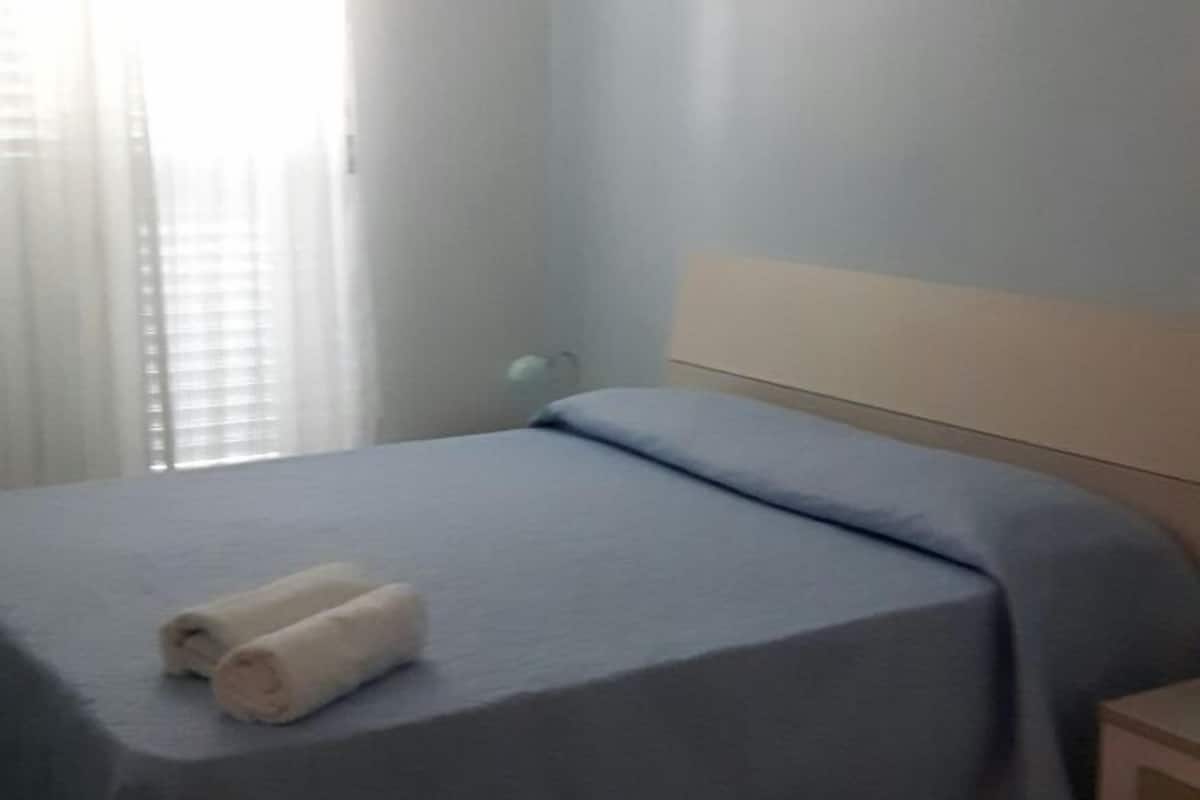 A bedroom with a bed, a nightstand, a lamp, and a window with blinds.