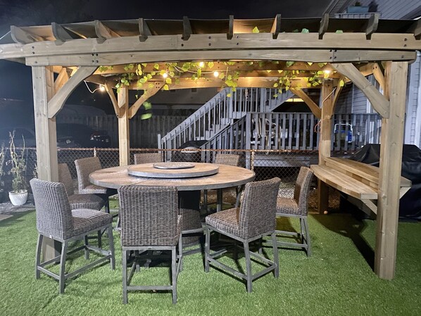 Una pérgola de madera con luces colgantes e hiedra, una mesa redonda con sillas y un área exterior cercada.