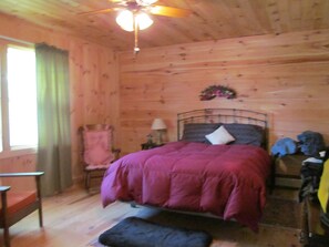 A wooden cabin bedroom with a bed, a chair, a nightstand, and a ceiling fan. - Comfy Home in the Adirondacks, close to Gore Mnt, hiking & snowmoble trails (Indian Lake)