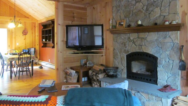Un interior de madera con chimenea de piedra, un televisor montado en la pared y un comedor con sillas y una mesa.