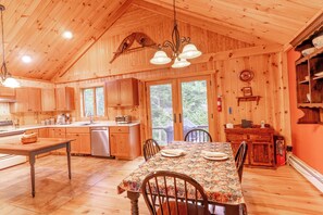 A wooden cabin interior with a dining table, chairs, a kitchen area, and a large window.