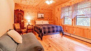 Habitación tipo cabaña de madera con una cama, una silla, una mesita de noche, un ventilador de techo y una ventana con cortinas de encaje.