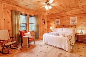A wooden cabin bedroom with a bed, two bedside lamps, a rocking chair, a window with curtains, and a ceiling fan. - Comfy Home in the Adirondacks, close to Gore Mnt, hiking & snowmoble trails (Indian Lake)