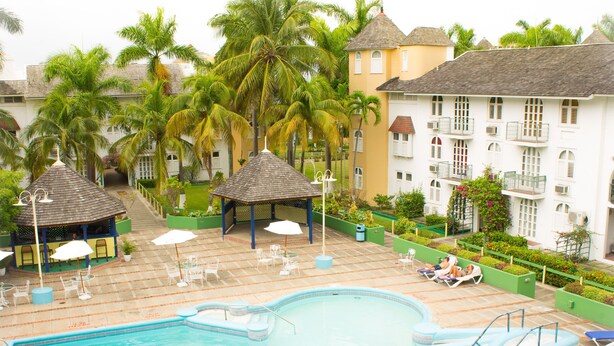 Un espace piscine agrémenté de chaises longues et de parasols, entouré de palmiers et bordé d’un bâtiment doté de balcons.