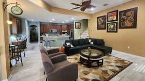 A spacious living area with a black sectional sofa, a coffee table, and a patterned rug. A kitchen with wooden cabinets and a dining area with a table and chairs are visible in the background.