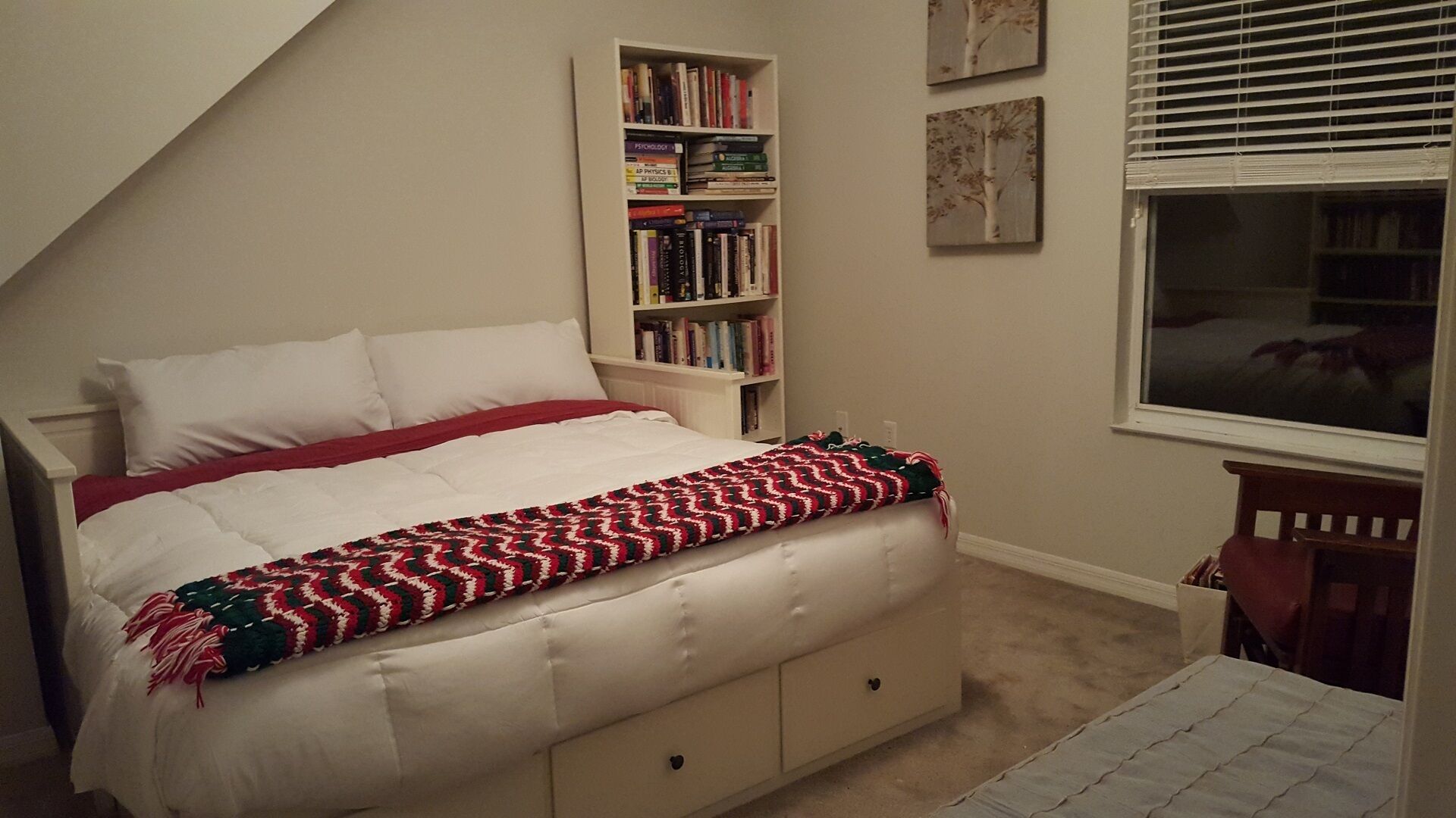 A bedroom with a bed, a bookshelf, a window, and a small table.