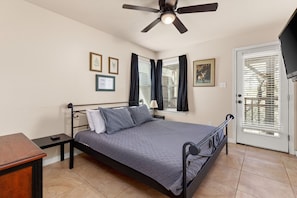 A bedroom with a bed, a nightstand, a desk, a ceiling fan, and a door with blinds.