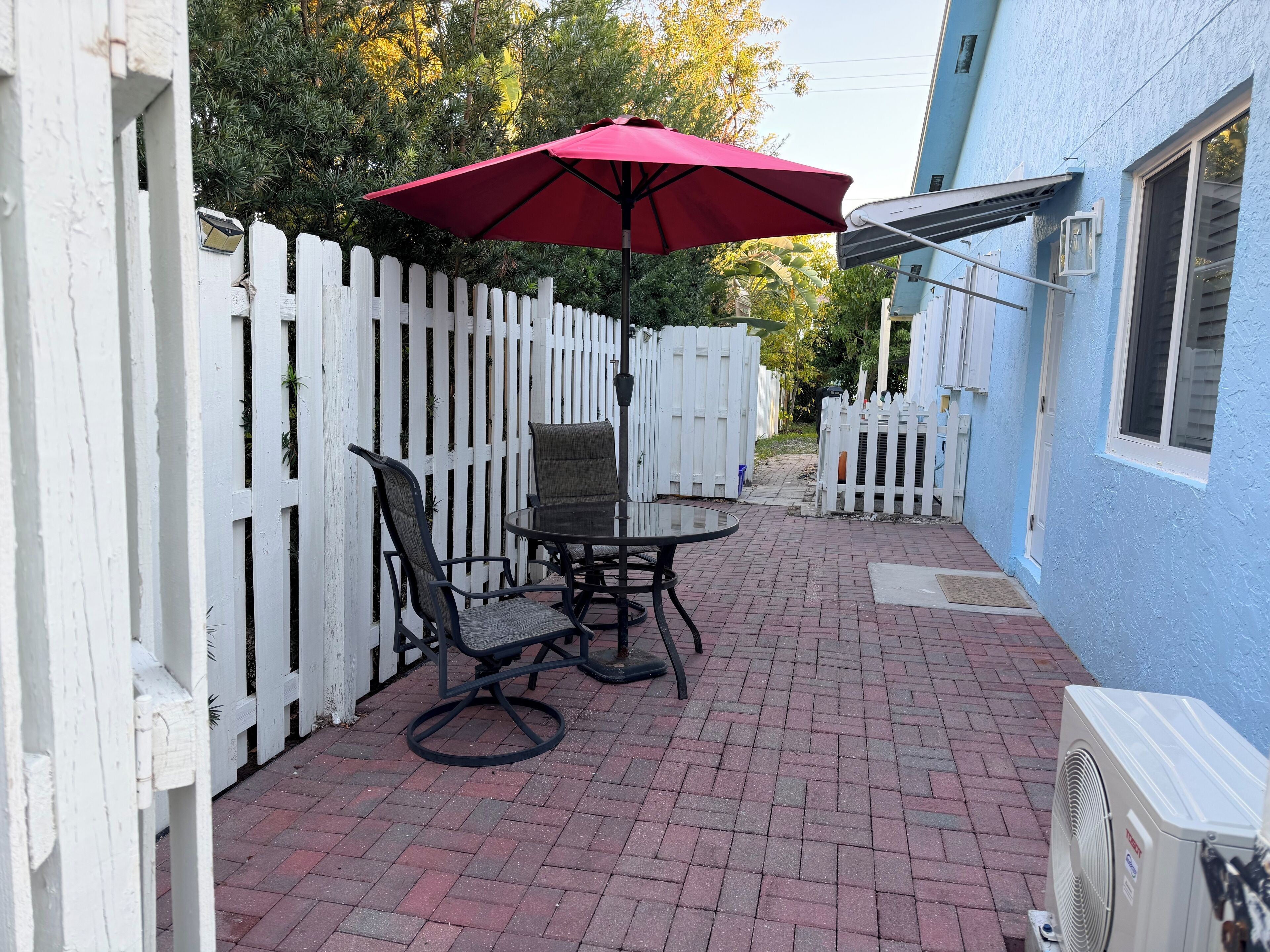 A patio with a table and chairs, a red umbrella, and a white picket fence.