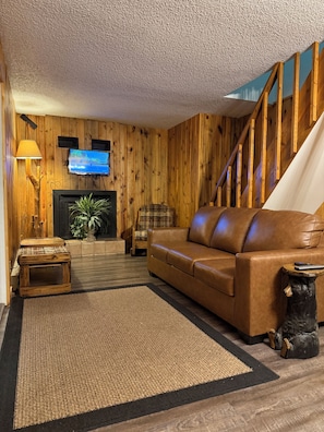 Una sala de estar acogedora con una pared de madera, un sofá de cuero, una chimenea, un televisor y una escalera.