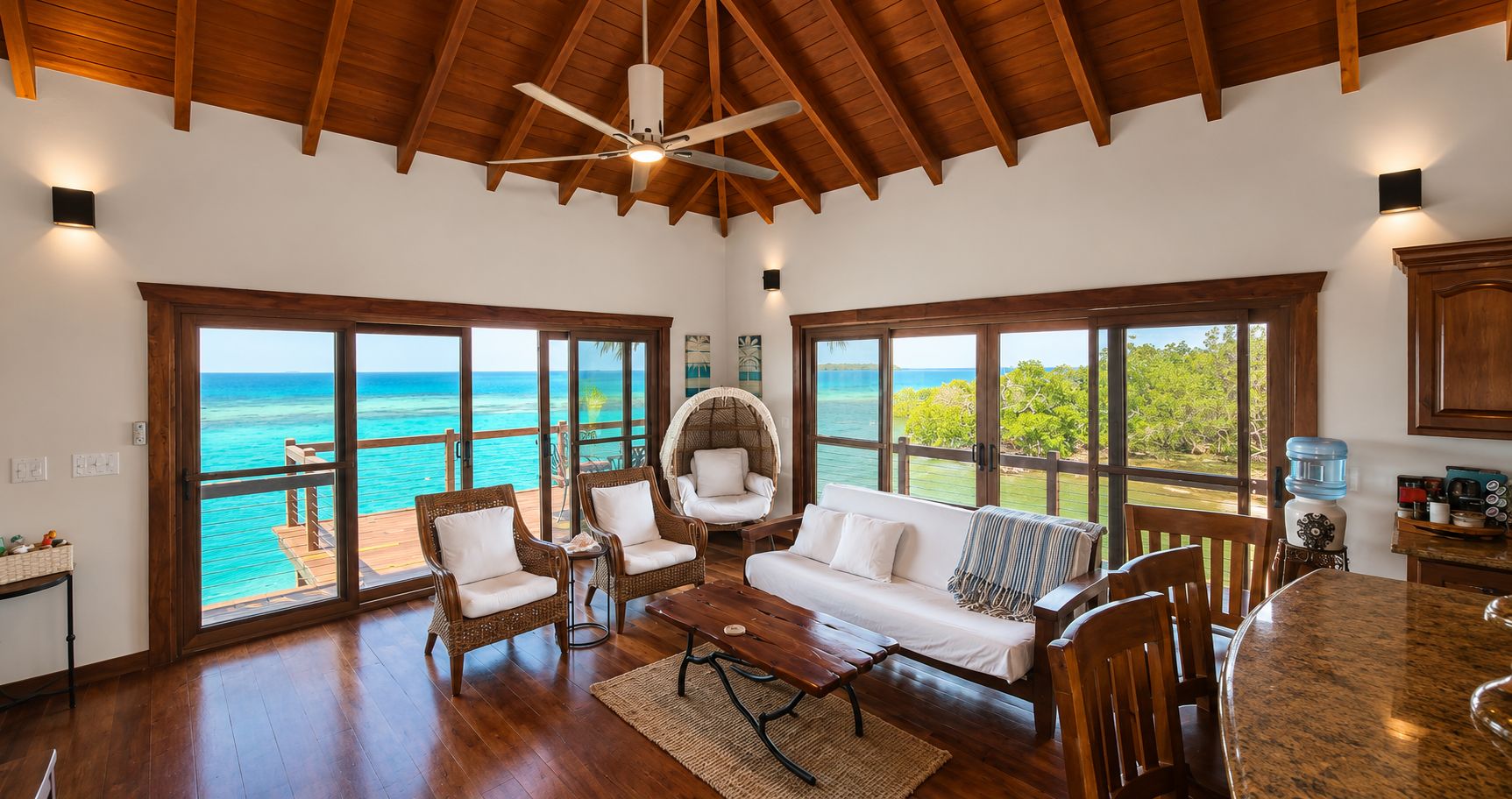 A spacious room with a wooden ceiling, large windows offering a view of the ocean, a dining area with a round table, and a seating area with wicker furniture.
