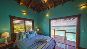 A bedroom with a bed, a nightstand, a lamp, and a view of the ocean through a large window. - All INCLUSIVE BELIZE PRIVATE ISLAND RENTAL WITH OVER WATER BUNGALOW AND CHEF (Belize City)