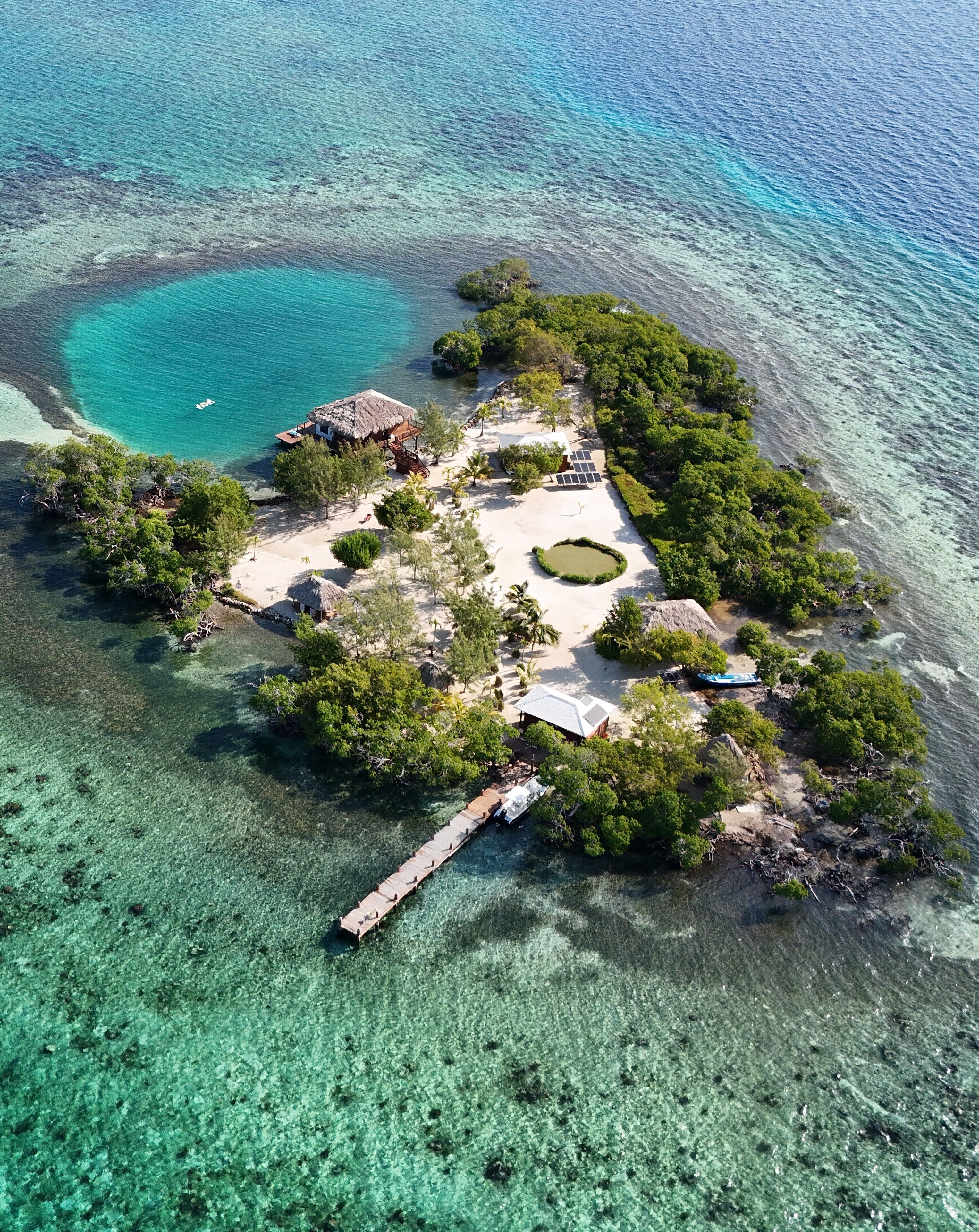 Une vue aérienne d’une île tropicale aux eaux turquoise et limpides, avec une jetée en bois et une zone centrale comprenant une structure à toit de chaume et un bâtiment blanc.
