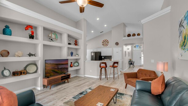 A modern living room with a TV, sofas, a coffee table, and a kitchen in the background. - Ashland Palms: Modern pool home, close to Disney on Glenbrook Resort  (Clermont)