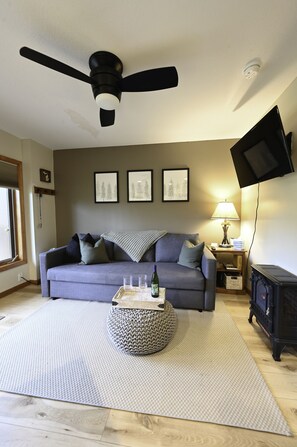 A living room with a grey sofa, a round ottoman, a ceiling fan, a fireplace, and a mounted TV. - 1 bedroom sleeps 4. On the Schuss golf course. close to Torch Lake and others. (Mancelona)