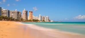 A beachfront hotel with multiple high-rise buildings, a sandy beach, and clear blue waters. - PentHouse Ocean views, Top-Roof Pool,10 Steps to Beach, Groups, max.9 2bed/2bath (San Juan)
