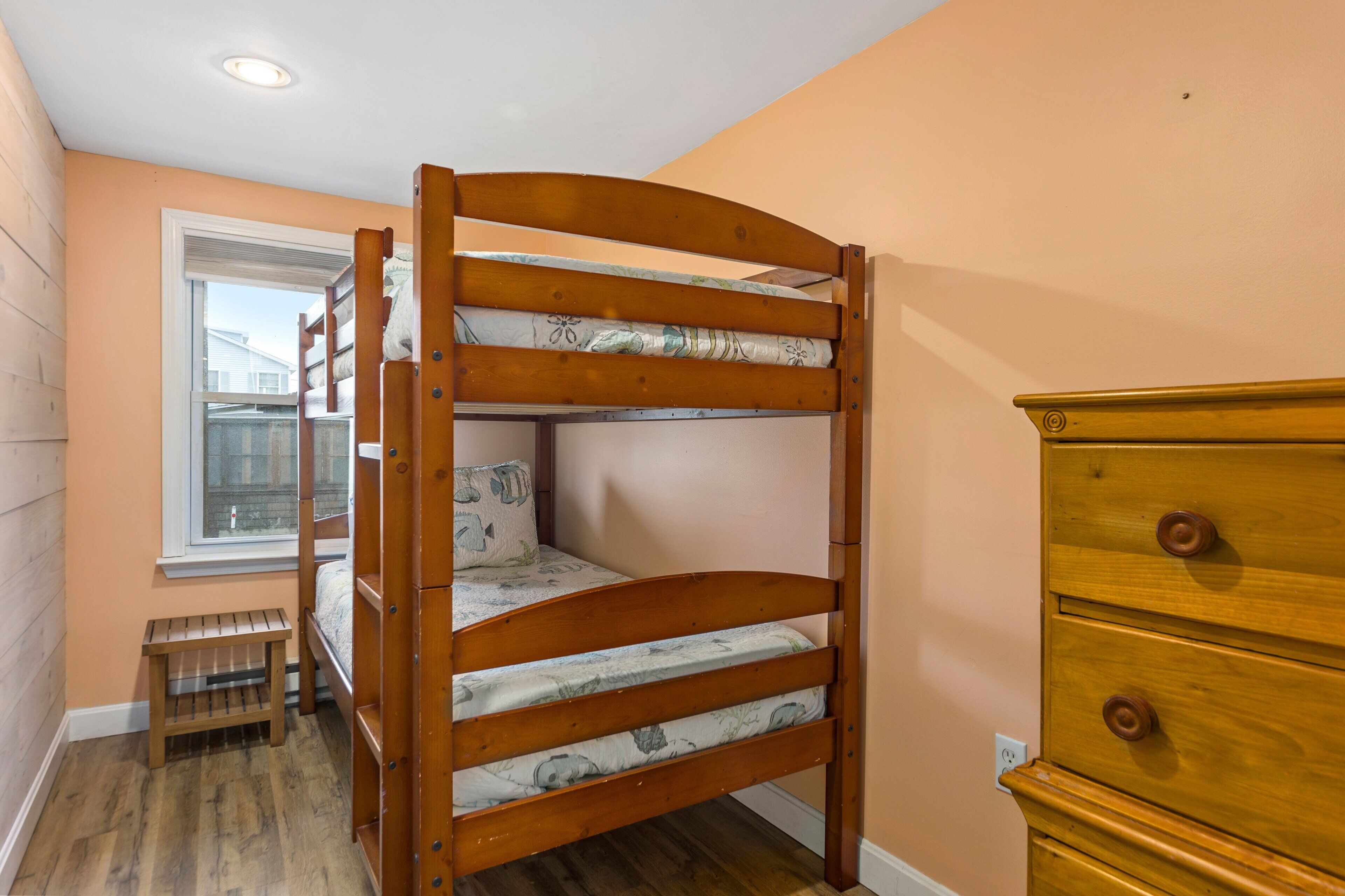 Un lit superposé avec une structure en bois et une literie à motifs, une commode en bois et une fenêtre avec des stores.