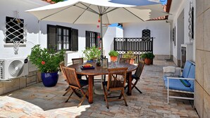 A patio with a round wooden table, chairs, a white umbrella, potted plants, and a white wall with a window and shutters.