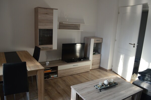 Un espace de vie compact comprenant une table à manger en bois, un téléviseur posé sur un meuble, un petit meuble de rangement et une table basse avec un plateau.
