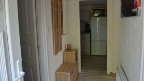 A narrow hallway with wooden cabinets and a small table. A kitchen with a refrigerator and microwave is visible in the background.