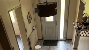 Un corridoio con piastrelle a motivi, una porta bianca, uno sgabello bianco, un cesto di vimini e un lampadario.