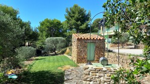 Un bâtiment en pierre avec une porte verte, entouré d’un jardin bien entretenu où poussent différentes espèces d’arbustes et d’arbres.