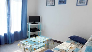 A room with a television on a blue stand, a table with a fish pattern tablecloth, a bed with a blue pillow, and a window with blue curtains.