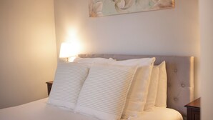 Un lit bien fait, agrémenté d’oreillers à rayures et d’une tête de lit capitonnée, une lampe de chevet et une œuvre d’art florale accrochée au mur.