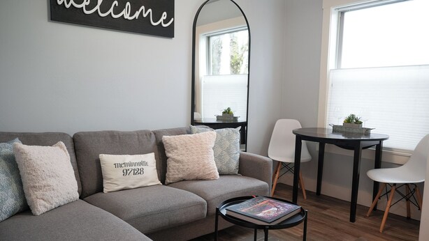 Un salon moderne avec un canapé d’angle gris, une petite table ronde sur laquelle repose un livre, et un grand miroir accroché au mur.
