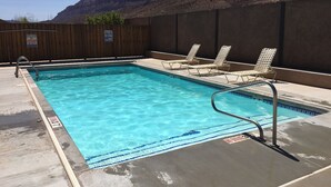 A rectangular swimming pool with clear blue water, surrounded by a wooden fence and a concrete deck. There are lounge chairs along the poolside, and a rocky mountain backdrop. - Westwater - Spacious 3 Bed, 2.5 Bath Moab House - Sleeps 8 (Moab)