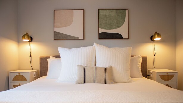 A neatly made bed with white bedding and multiple pillows, flanked by two bedside tables with lamps, in a room with abstract artwork on the walls.