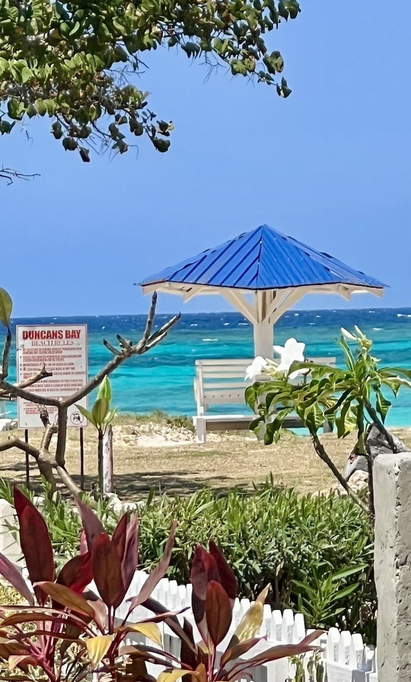 A beach with a blue umbrella, a bench, and a sign for Duncan's Bay. - ONE RHYTHM VILLA - Steps to the BEACH - CHEF Included - Caribbean Sea Views (duncans bay)