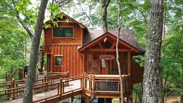 Une cabane en bois perchée dans un arbre, dotée d’une terrasse en bois et d’une pancarte indiquant : « Cabane dans les arbres du terrain de camping ».