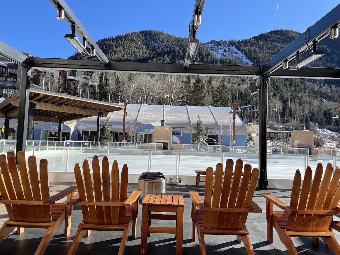 A covered outdoor seating area with wooden chairs and tables, overlooking a ski resort with buildings and mountains in the background.