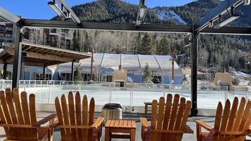 A covered outdoor seating area with wooden chairs and tables, overlooking a ski resort with buildings and mountains in the background.
