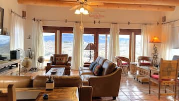 A spacious living room with a high ceiling, leather sofas, a wooden coffee table, and a television. Large windows offer a view of the landscape.
