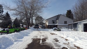 A residential area with snow-covered ground, a house with a garage, and several snowmobiles parked in the driveway.