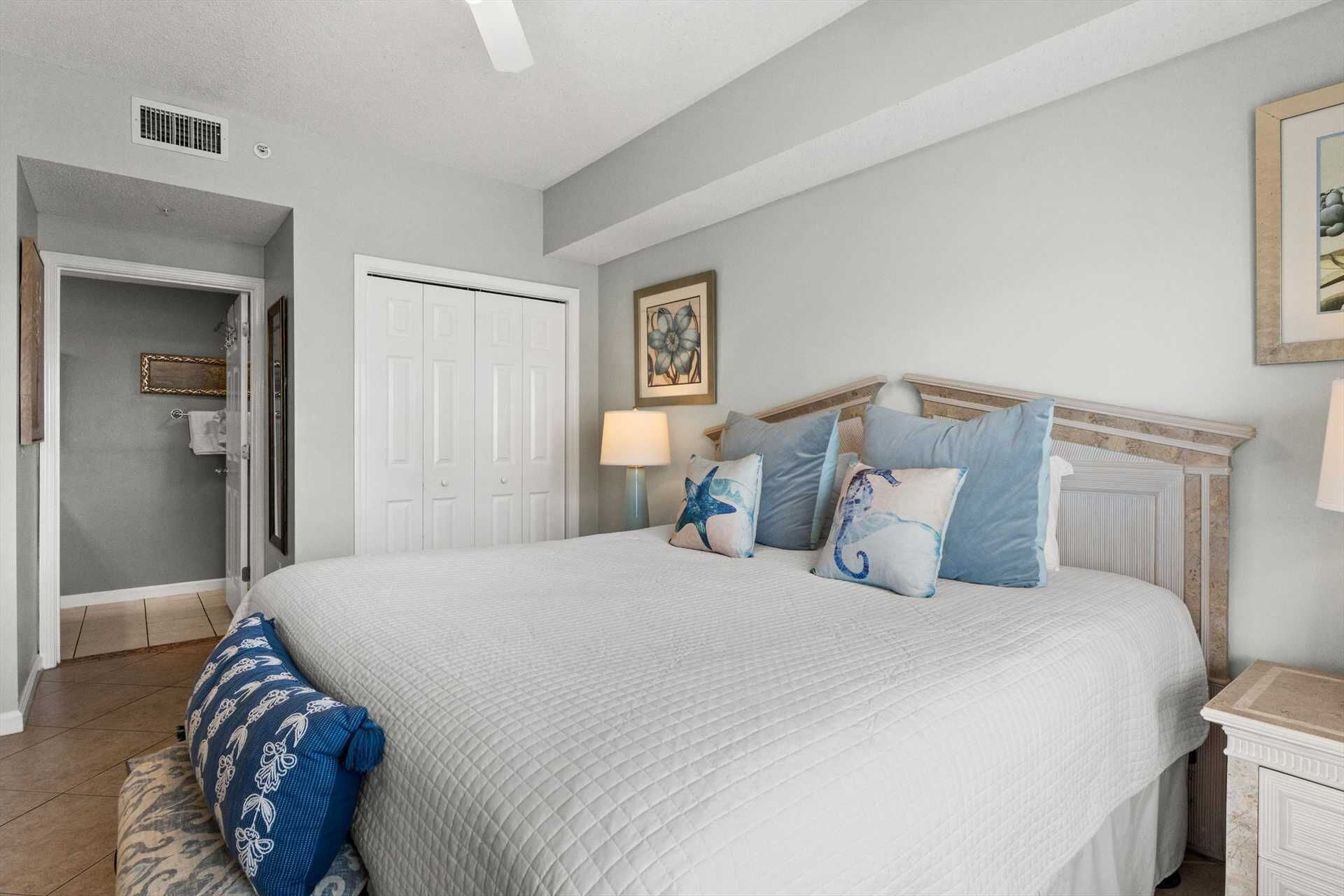 A bedroom with a bed, a nightstand, a lamp, a framed picture on the wall, and a door leading to another room.