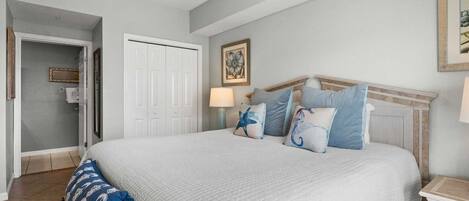 A bedroom with a bed, a nightstand, a lamp, a framed picture on the wall, and a door leading to another room.