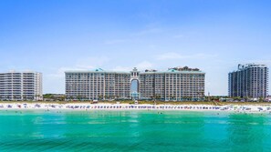 Un hotel frente a la playa con varios edificios altos, un arco central y una playa de arena con sombrillas.